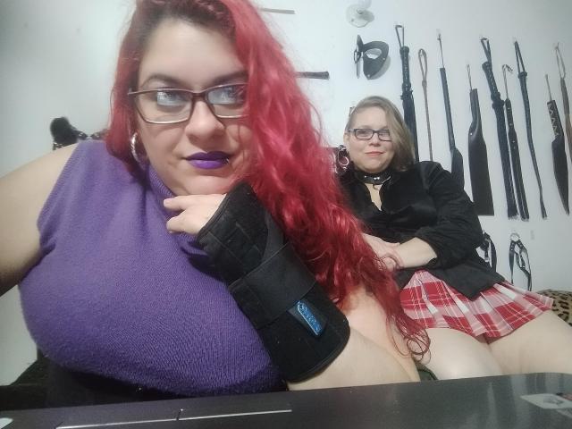 Duas latinas com óculos, uma com cabelo ruivo e seios grandes vestindo uma blusa preta, e a outra com cabelo loiro usando um corpete roxo, em pé em um quarto..