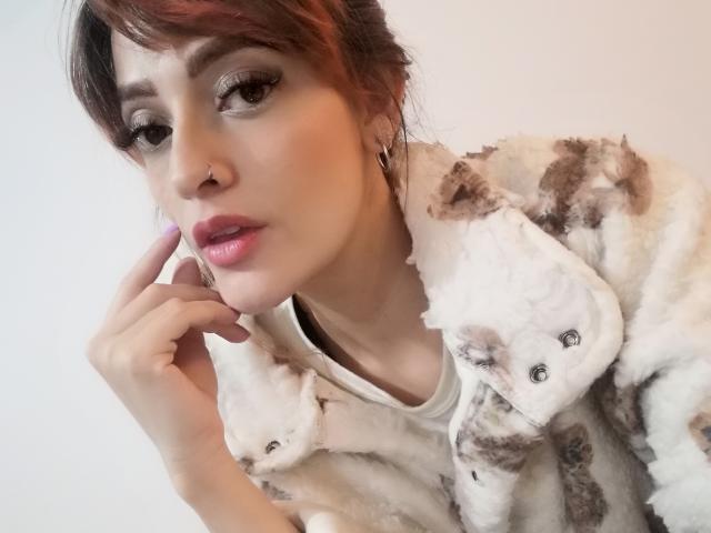 JaneRoa with brown hair, nose ring, and pink lips, wearing a fluffy white and brown coat with a cat face, touching her cheek, looking at the camera.