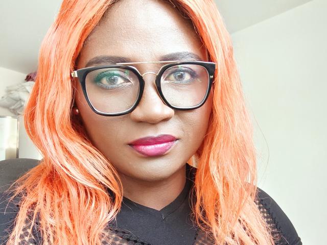SkyleurX a les cheveux orange, des lunettes noires et du rouge à lèvres rose. Elle porte une chemise noire avec des détails en dentelle. Ses yeux sont verts, et elle regarde directement l'appareil photo..