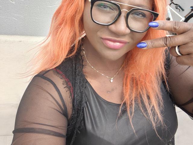SkyleurX com cabelo laranja, blusa preta transparente, unhas azuis, e óculos, mostrando um sinal de paz, sorrindo, lábios rosados, e uma pequena tatuagem no peito..