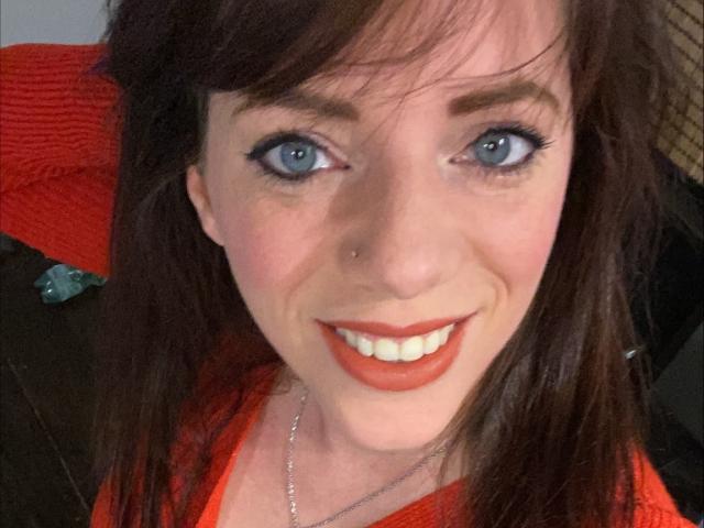 SerenaFr, con ojos azules, cabello castaño, lápiz labial rojo y un piercing en la nariz, sonríe a la cámara y lleva una blusa naranja. Sus senos son parcialmente visibles debajo de la tela..
