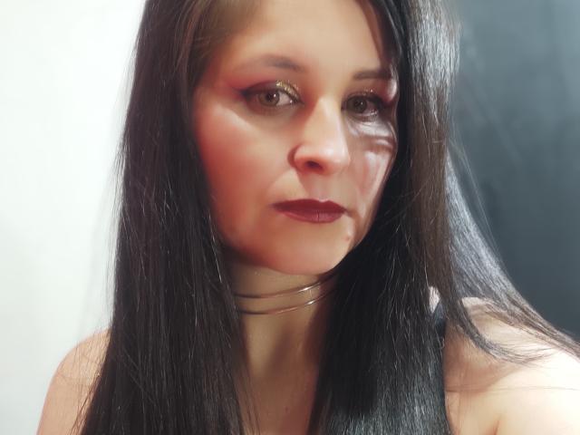 KynkyFetish has long black hair, shiny wet skin, dark red lipstick, and a metallic choker. She's looking slightly to the side with a subtle, confident expression.