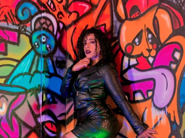 Scarlett, con i capelli neri ricci, indossa un abito di pelle nera lucida, in piedi di fronte a graffiti colorati, con un dito vicino alla bocca aperta, una luce verde sulla coscia..