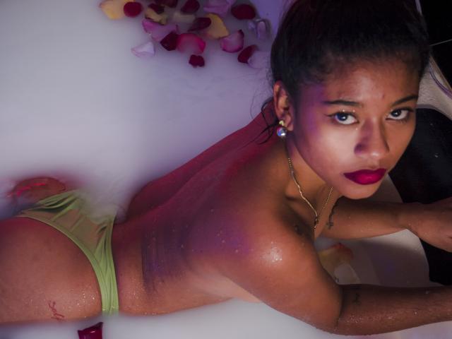 TamiDubois, con la pelle scura, sdraiata in una vasca da bagno, indossa delle mutandine verdi, con la schiena rivolta verso la telecamera, guarda oltre la spalla, rossetto rosso, orecchini, acqua, petali di rosa, pelle bagnata, sexy, attraente..