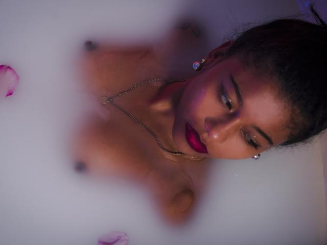Le visage de TamiDubois et ses seins nus dans un bain brumeux, avec des pétales roses, portant un collier et du rouge à lèvres, regardant vers le bas avec ses cheveux bruns foncés relevés..