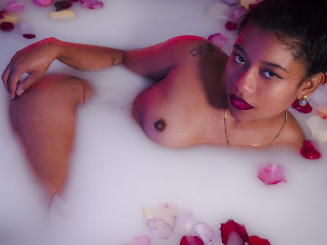 TamiDubois in einer Schaumbad, nackt mit Rosenblüten, dunklem Make-up und einer goldenen Halskette, ihre Brüste über dem Wasser, blickt mit einem verführerischen Ausdruck in die Kamera..