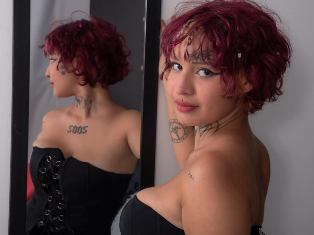 SayuriRose, com cabelos ruivos cacheados, um corpete preto sem alças e tatuagens, olhando para seu reflexo em um espelho, exibindo seus pequenos seios e um sorriso confiante..