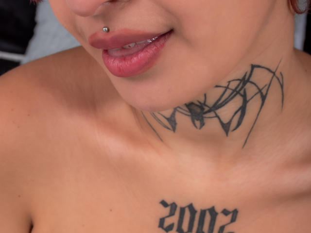 La parte superiore del corpo di SayuriRose è visibile, mostrando il suo piercing al labbro, le labbra rosa lucide, il collo tatuato e il petto tatuato con la scritta "2012" in caratteri grassetti. È senza top e ha la pelle abbronzata..
