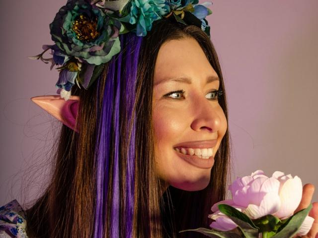 MisissFire with elf ears, purple and brown hair, flower crown, smiling, holding pink peony, soft pink background, wearing purple dress, cute and fantasy-like.