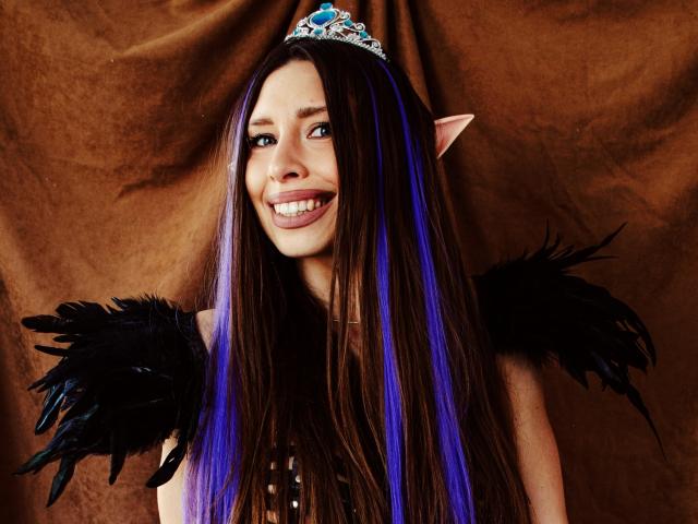 MisissFire with long brown and blue hair, elf ears, black feather wings, and a silver tiara, smiling with visible teeth. She's wearing a beaded top, looking cute.