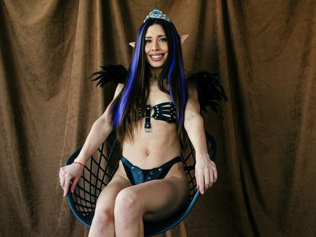 MisissFire with elf ears, blue and purple hair, black feather wings, black strappy bikini, and a tiara, sitting on a blue chair, smiling at the camera.