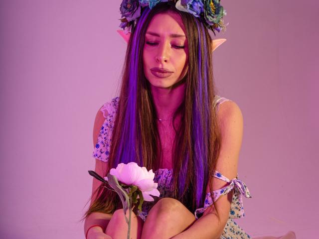 MisissFire with long brown hair, elf ears, flower crown, and floral dress holding a pink flower, sitting with eyes closed, purple and pink lighting on her skin.