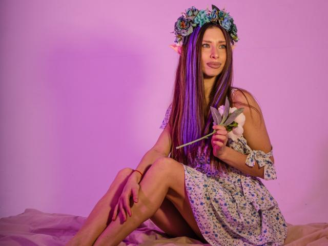 MisissFire with long purple and brown hair, wearing a floral dress and flower crown, holding a white lily, sitting on a pink-lit surface with legs crossed.