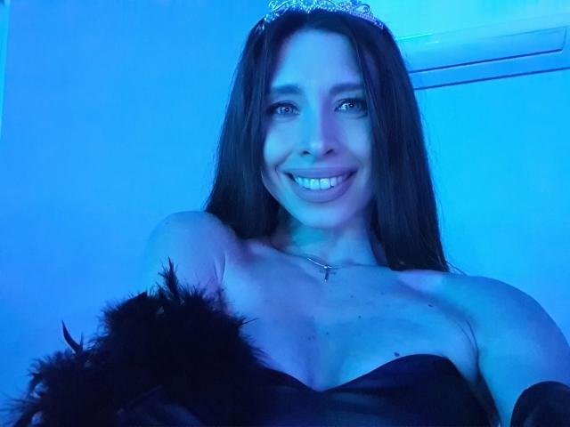 MisissFire with long brown hair, blue light, smiling, wearing a black strapless dress, feather accessory, and silver tiara, showing her tits and a small cross necklace.