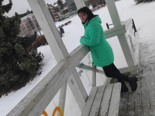 NikaMel, une femme corpulente aux cheveux noirs, portant une veste verte matelassée et un pantalon noir, est appuyée contre un pont en bois recouvert de graffitis, dans un décor enneigé et nuageux..