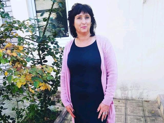 NikaMel a les cheveux noirs, un cardigan rose et une robe noire. Elle se tient debout avec un léger sourire, les mains le long du corps, à côté d'un buisson aux feuilles vertes et jaunes..