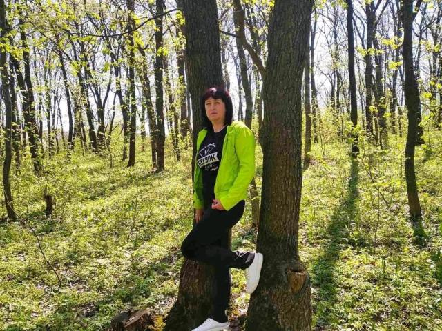 NikaMel with black hair, green jacket, black shirt, black pants, and white sneakers, leaning on a tree in a forest, legs crossed, looking at camera, daytime.