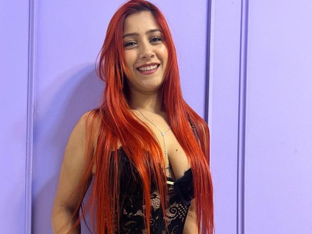 Lisa Harvie, con lunghi capelli rossi, sorridente, indossa un reggiseno di pizzo nero, mostrando il seno, in piedi di fronte a un muro viola, appare carina e sexy..