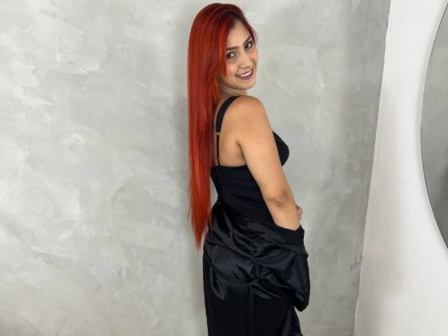LisaHarvie with long red hair, black dress, smiling, looking over shoulder, hands in pockets, standing against gray textured wall, right side of image shows white circular mirror.