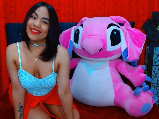 SharomCastle with pink baby angel doll, wearing blue lace top, red skirt, choker, smiling, showing small tits, red lipstick, black hair, sitting next to giant pink plushie.