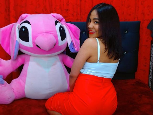 SharomCastle com um brinquedo de pelúcia rosa ao lado dela, vestindo uma camiseta branca e uma saia vermelha, sentada em uma cama vermelha, sorrindo, mostrando o bumbum, cabelo preto, batom vermelho, fofa..