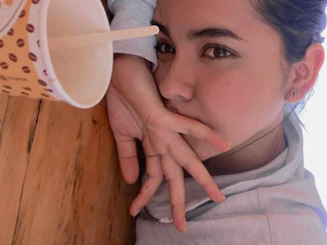 O rosto de Liana Bernocchi está quase submerso em uma xícara de café, e sua mão cobre a boca. Ela está vestindo um moletom cinza, com o cabelo castanho preso em um coque..