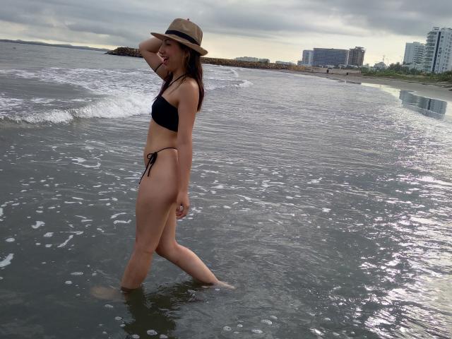 EmaParis, standing in shallow water, wears a black bikini and a beige hat, with her hand on her head, showing her ass and small tits. Overcast sky.