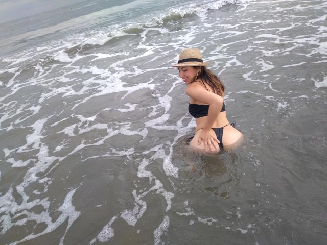 EmaParis usando um biquíni preto, um chapéu de cor clara e cabelo castanho molhado, sorrindo enquanto está de joelhos em águas rasas do oceano, com a bunda e a vulva parcialmente visíveis..