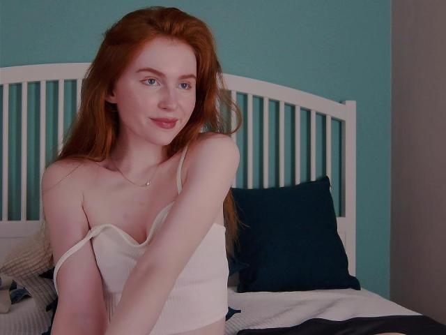 GingerArina mit roten Haaren, heller Haut und kleinen Brüsten, trägt ein weißes Tanktop, das sie leicht herunterzieht, und lächelt. Sie liegt auf einem Bett mit blau-weißen Kissen..