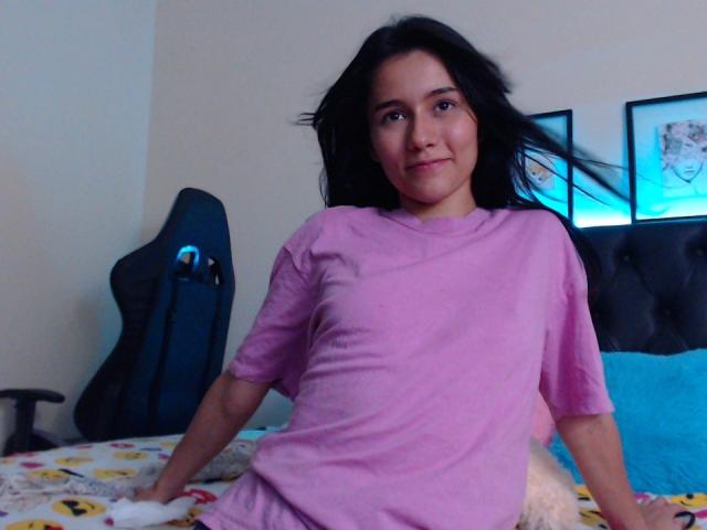 DeisyWest with black hair, pink shirt, and slight smile, sitting on bed with blue pillow, black chair in background, and framed pictures on wall. Her tits are small.