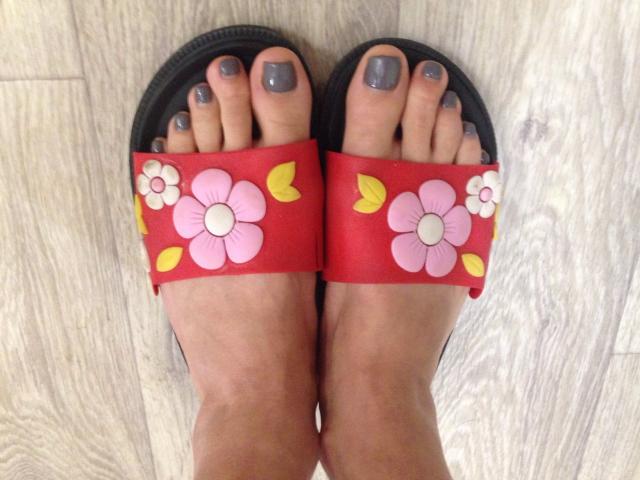 AnabelSex69's feet with gray-painted toenails, wearing black flip-flops with red straps and pink flower decorations, standing on a wooden floor.