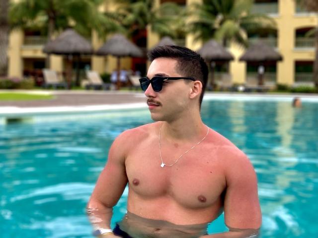 Andosia, shirtless, in a pool, wearing black sunglasses, with a mustache, short dark hair, and a silver necklace, showing a toned, red-skinned chest, standing in clear water.