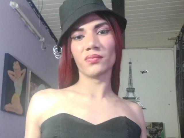 LunaGransTs with red hair, black hat, black strapless top, pink eyeliner, and pink lipstick, showing a slight smile, with a black ring in her right ear.