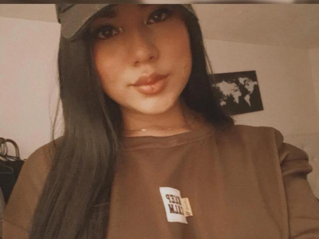 SteffanyTaylor with long black hair, brown eyes, and full lips, wearing a brown cap and brown sweatshirt, showing her pouty ass-kissing face. Warm, slightly blurry selfie.