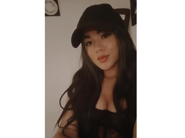 SteffanyTaylor with long black hair, wearing a black cap and a revealing black top showing her tits, has red lipstick and a cute smile, looking at the camera.