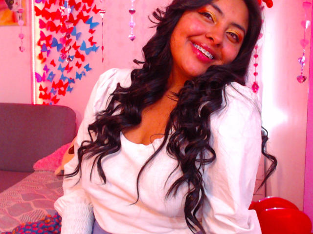 AngelaRoggers, com cabelo cacheado escuro, sorrindo, vestindo uma camisa branca, mostrando os seios, batom rosa, maquiagem amarela nos olhos e lábios vermelhos, sentada em um quarto rosa..