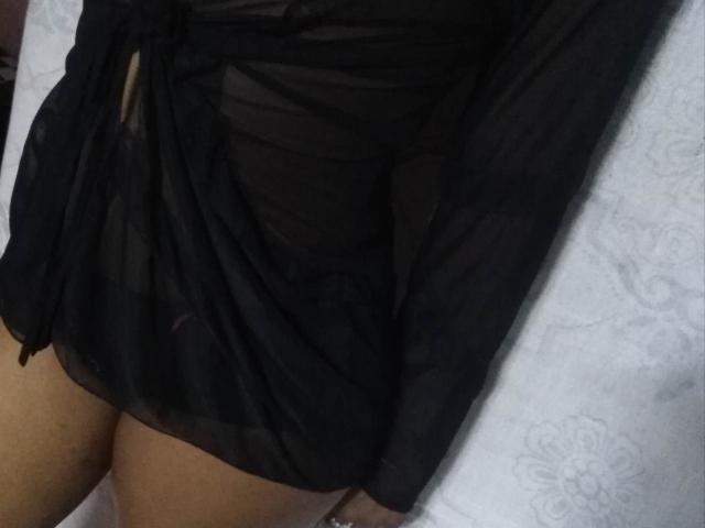 O vestido preto, transparente e de mangas compridas de OliviaBliss está levantado, expondo suas coxas bronzeadas e uma pequena fenda visível no tecido, revelando um vislumbre de sua vulva..