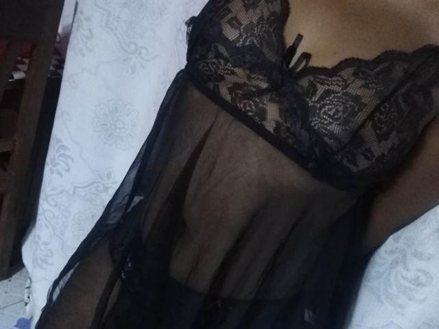 OliviaBliss in biancheria intima di pizzo nero con tessuto trasparente, che mostra il seno e un fiocco nero sulla parte superiore, sdraiata su un letto bianco con motivi..