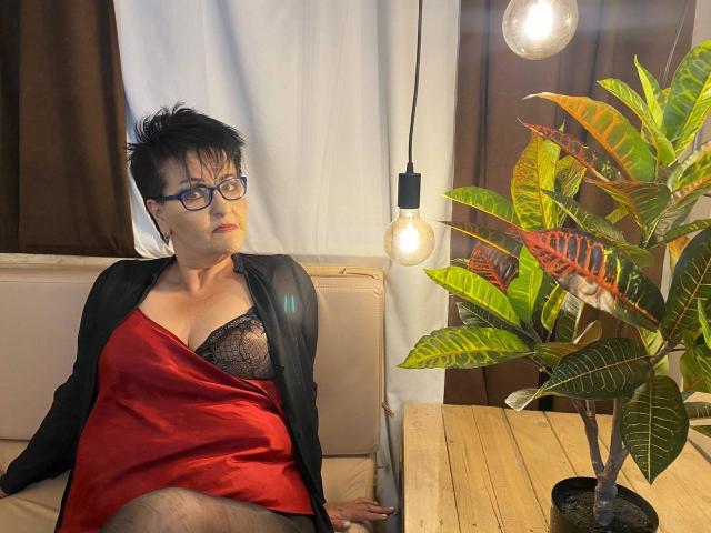 SondraRiddle with short black hair, glasses, red satin dress, black lace bra, black robe, sitting on beige sofa, next to potted plant and exposed bulb, wooden table.