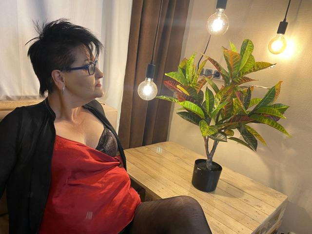 Sondra Riddle, mit kurzen schwarzen Haaren, Brille und einem roten Hemd, das ihre schwarzen, mit Spitze bedeckten Brüste freilegt, sitzt neben einem Topf mit einer Pflanze mit hängenden Glühbirnen in einem Raum..