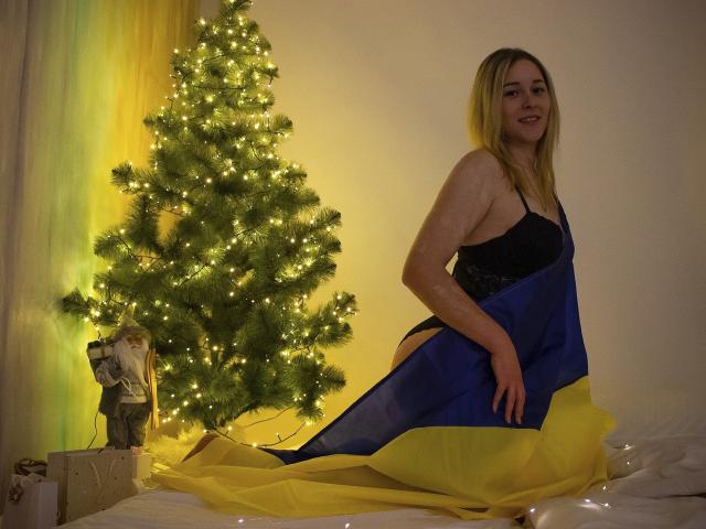 DoraSevi with blonde hair, black lingerie, and blue-yellow fabric, sitting on bed by lit Christmas tree, smiling, showing ass, with small statue and gift on left.