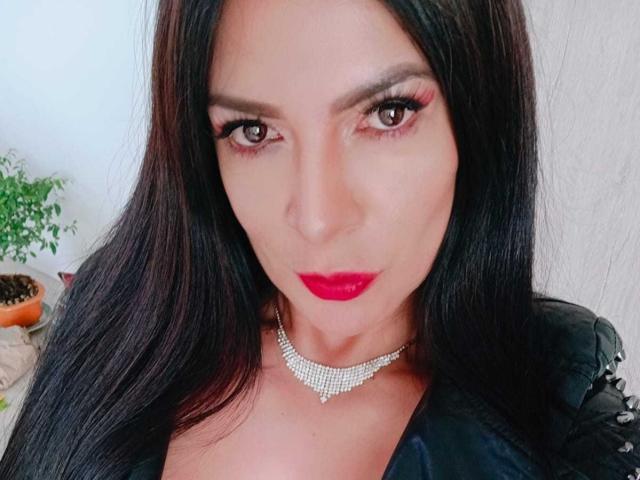 JoTaylor tiene el cabello oscuro y lacio, lápiz labial rojo y un collar brillante. Está mirando a la cámara con ojos oscuros y expresivos. Se pueden ver los senos debajo de una blusa negra..