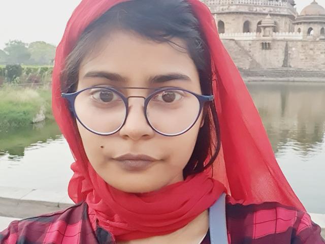 Sara Mehta, usando óculos, um lenço vermelho na cabeça e uma camisa xadrez, mostrando uma pequena marca de beleza na bochecha direita, está em pé perto de uma fonte de água, com um edifício histórico ao fundo..