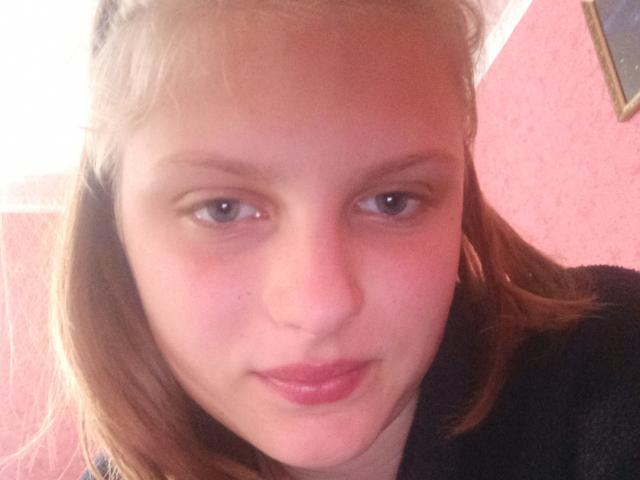 KoketaOrrie a un visage mignon avec des cheveux bruns clairs, de petits yeux marrons et des lèvres rosées. Elle porte un haut noir, a un léger sourire et a une peau claire..
