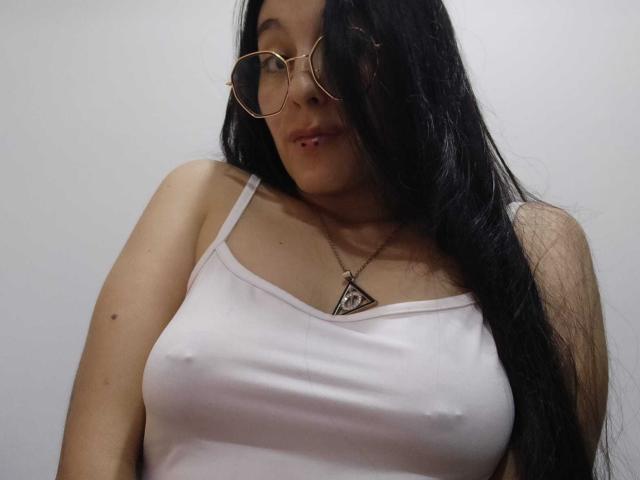 LatinAleja, com cabelos longos e pretos, usando óculos e uma camiseta branca que mostra seus mamilos. Ela tem um colar em formato de triângulo e uma expressão levemente emburrada..