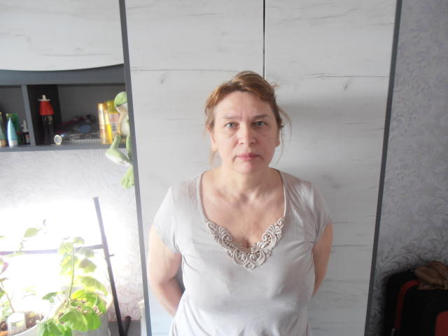AlineLoveFlowers, een vrouw van middelbare leeftijd met lichte huid en bruin haar in een paardenstaart, draagt een grijze top met een decoratieve halslijn en staat met haar armen achter haar rug, met een serieuze blik..