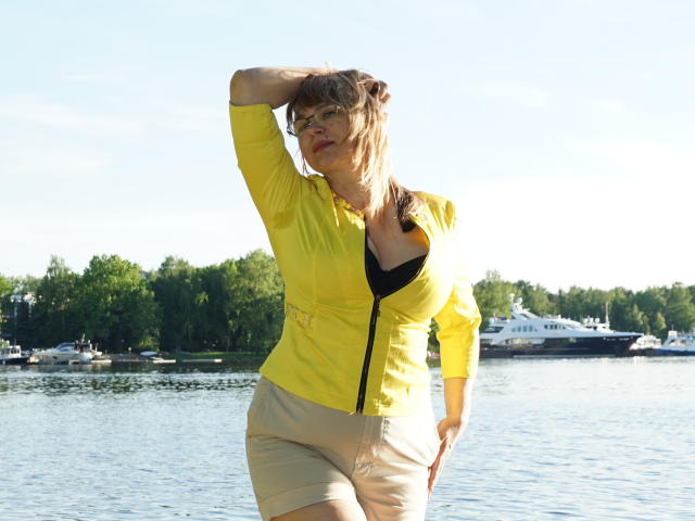 AlineLoveFlowers steht am See, trägt ein gelbes, vorne zuziehendes Hemd, unter dem ihr schwarzer BH und beige Shorts zu sehen sind, und hält ihre Hand in ihrem braunen Haar, sonniger Tag..