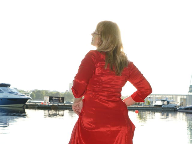 AlineLoveFlowers está de costas para a câmera, vestindo um vestido vermelho brilhante, com as mãos nos quadris, a luz do sol iluminando seus cabelos loiros, ao lado de um barco atracado..