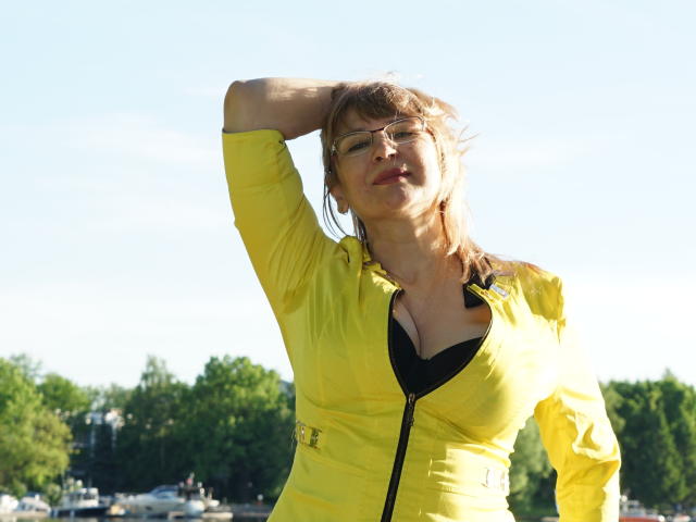 AlineLoveFlowers, com cabelo loiro, óculos e um casaco amarelo, com a mão no cabelo, mostrando seu sutiã preto e seios médios, em pé ao ar livre sob a luz do sol..