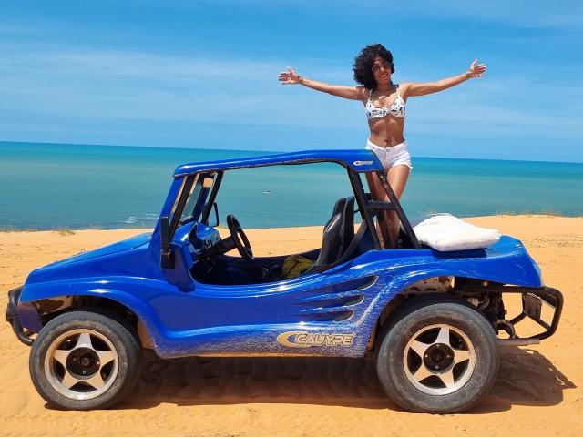 LolaPetterson with curly black hair, wearing a white bikini, stands with arms out on a blue G-Force buggy, on golden sand, near clear blue ocean.